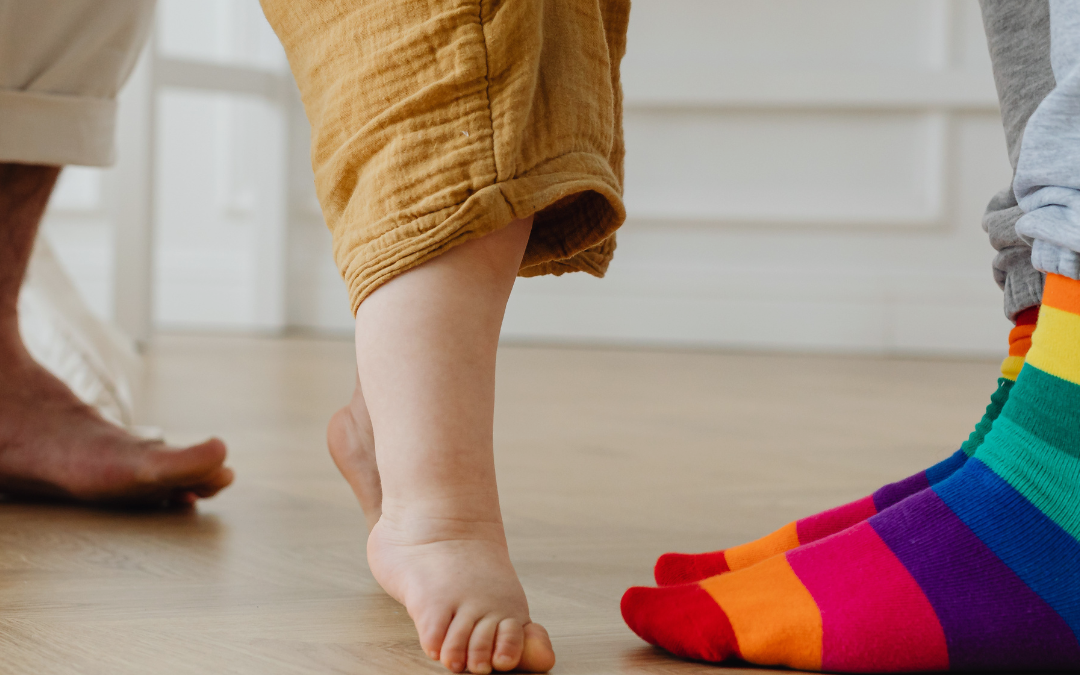 Two adults stand on either side of a toddler, holding the child’s hands as they learn to walk. One adult is wearing rainbow-striped socks.