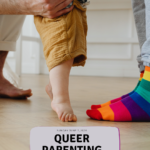 Two adults stand on either side of a toddler, holding the child’s hands as they learn to walk. One adult is wearing rainbow-striped socks.